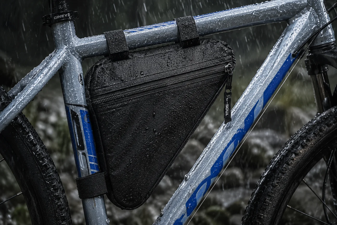 Black triangle frame bag strapped inside the frame of a silver and blue mountain bike during heavy rain, water beading on the fabric and zipper