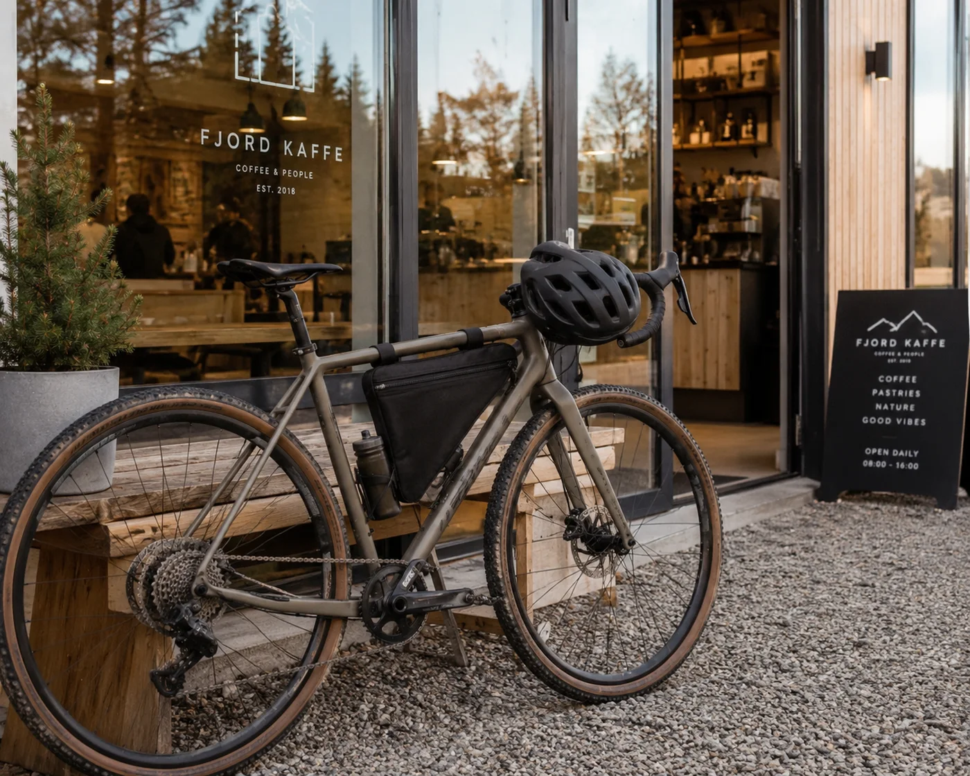 Sand-coloured gravel bike parked outside a coffee shop with a black triangle bag high in the frame and a water bottle in the down-tube cage just below it